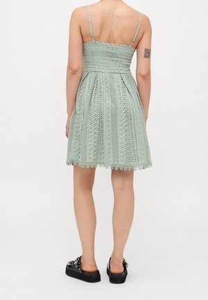 Femme portant une robe en dentelle vert menthe à fines bretelles, chaussures à plateforme noires sans lacets, et un tatouage floral noir sur le haut du bras gauche.