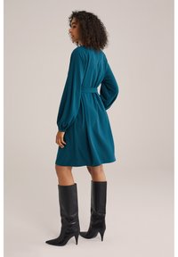 Teal klänning med långa ärmar och en figursydd midja, parat med knähöga svarta läderstövlar med spetsig tå, som visar en slät textur och enkel design.