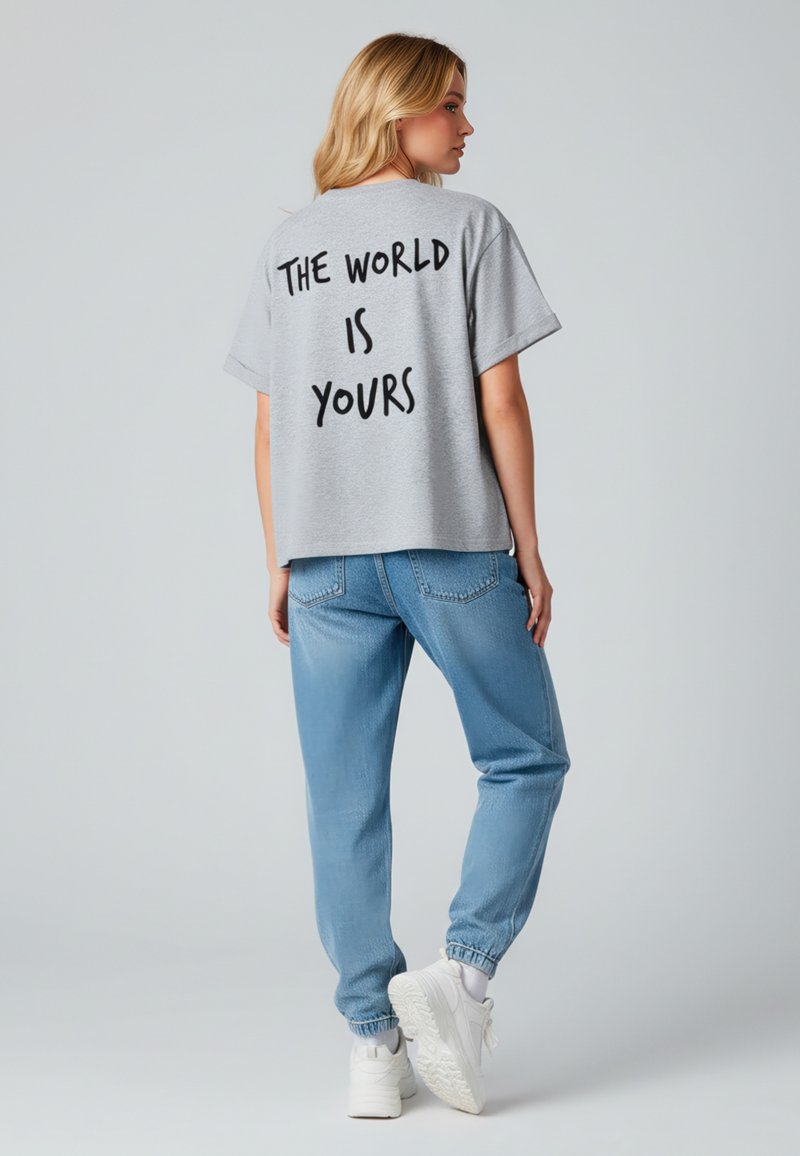 T-shirt gris surdimensionné avec des lettres noires au dos, associé à un jean jogger bleu clair et des baskets blanches. Tenue décontractée avec une coupe ample.