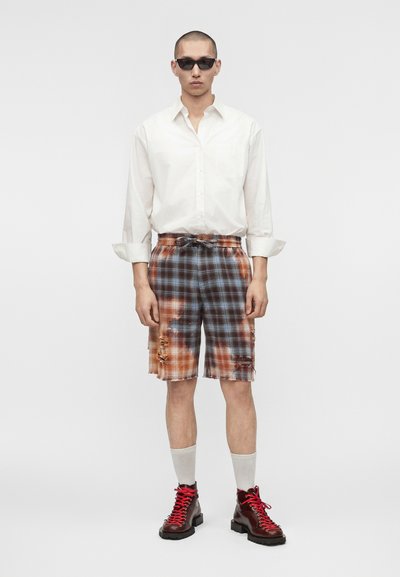 Άνδρας που φοράει μαύρα γυαλιά ηλίου, λευκό πουκάμισο με κουμπιά, βερμούδα καρό σε στιλ tie-dye με φθαρμένες λεπτομέρειες, λευκές κάλτσες και σκούρα κόκκινα μποτάκια.