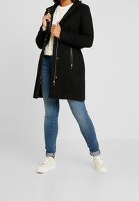 Manteau noir mi-long jusqu'aux genoux avec capuche, fabriqué en tissu doux. Comprend des poches zippées et une fermeture éclair lisse. Associé à un pull léger et un jean.