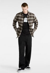 Karierter Flanellhemd in Braun und Beige, schwarzes Grafik-T-Shirt, lockere schwarze Hose und schwarze Sneakers mit weißen Akzenten.