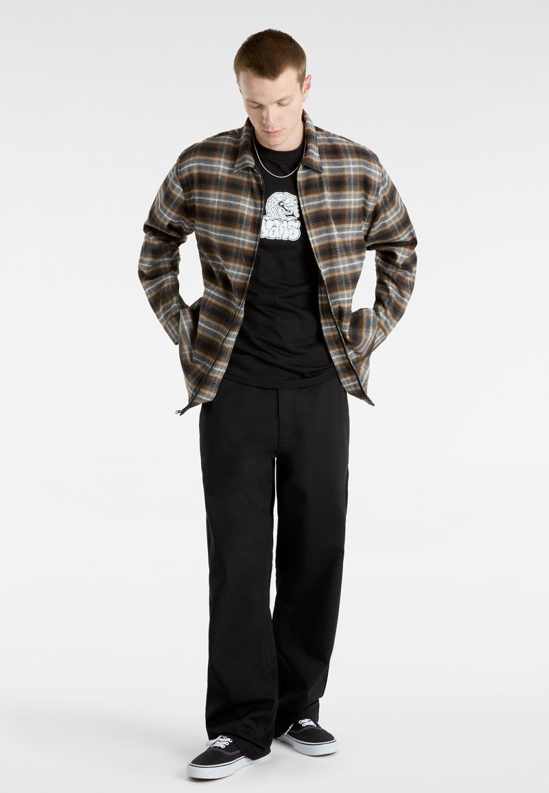 Karierter Flanellhemd in Braun und Beige, schwarzes Grafik-T-Shirt, lockere schwarze Hose und schwarze Sneakers mit weißen Akzenten.