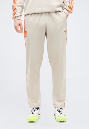 Pantalon de survêtement beige avec des rayures et des logos orange sur les côtés, porté avec des chaussures à crampons blanches dotées de semelles vert néon.