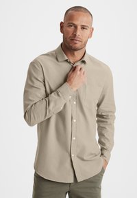 Beige Langarmhemd mit Knöpfen, einer Brusttasche, weißen Knöpfen und einer glatten Textur, kombiniert mit olivgrünen Hosen.