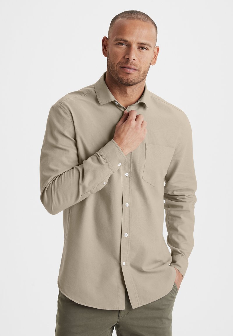 Beige Langarmhemd mit Knöpfen, einer Brusttasche, weißen Knöpfen und einer glatten Textur, kombiniert mit olivgrünen Hosen.