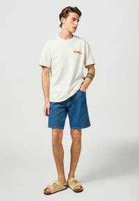 Lichtcreme t-shirt met "WRANGLER" logo, blauwe denimshorts en tan sandalen. Focus op casual streetwear esthetiek en een relaxte pasvorm.