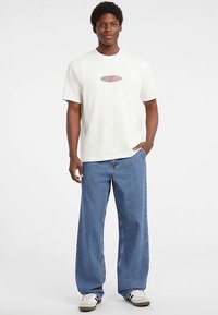 Fehér kerek nyakú póló kis ovális grafikával és kék bő szárú farmernadrág. A szettet casual lábbeli egészíti ki fekete-fehér részletekkel.