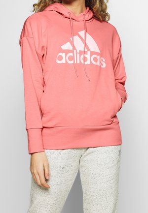 Person iført en pink Adidas-hoodie med hvidt logo og lysegrå joggingbukser, stående med den ene hånd i lommen mod en ensfarvet baggrund.