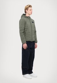 Khaki quilted jacket with a hood and black chest pocket, paired with dark blue wide-leg pants and white sneakers.
