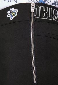 Pantalon noir avec une fermeture éclair argentée et un patch abeille brodé. Il présente un tissu lisse et extensible ainsi qu'une taille avec un logo.