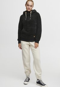 Un sudadera de felpa negra con capucha, que cuenta con un bolsillo frontal, cordones ajustables y puños de canalé, combinada con pantalones de chándal crema y zapatillas negras.