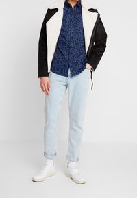 Czarna kurtka z czarnego imitacji zamszu z białą podszewką z polaru, niebieska koszula na guziki w cętki lamparta, jasnoniebieskie dżinsy i białe trampki.