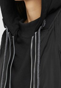 Czarna kurtka z kapturem, z szarym sznurkiem, wysokim kołnierzem i metalowymi zamkami błyskawicznymi z przodu. Szczegóły z teksturowanej tkaniny.