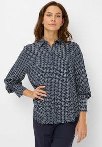 Vrouw in een marineblauwe, gemerkte blouse en donkere broeken, staande met één hand op haar heup tegen een effen achtergrond.