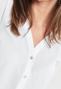 Blusa bianca realizzata in un tessuto leggero, caratterizzata da un design con scollatura a V, chiusura con bottoni e una texture liscia.