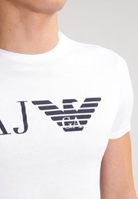 Vit kortärmad T-shirt med en stor svartrandig örnlogotyp tryckt på framsidan, d är bokstäverna "GA" är integrerade i designen.