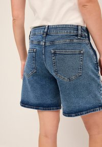 Pantalones cortos de mezclilla de un lavado azul medio con un diseño clásico de cinco bolsillos, dobladillos deshilachados y un ajuste relajado. Un ligero desgastado añade textura.