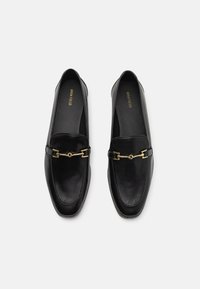 Mocassins en cuir noir avec un accent de chaîne en ton doré, semelle plate, bout arrondi et texture lisse. Design classique avec un minimum de détails.