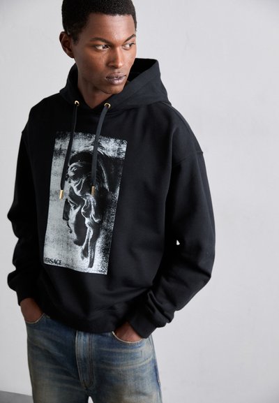 Sudadera negra con un gráfico texturizado de una figura clásica; cuenta con herrajes en tono dorado y un cordón ajustable. Combinada con unos jeans de mezclilla azul.