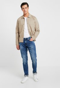 Jasno beżowa kurtka zapinana na guziki na białym t-shircie, w połączeniu z niebieskimi dżinsami i białymi trampkami. Gładki materiał o luźnym kroju.