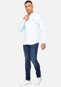 Camisa de botones azul claro con mangas largas, con un pequeño emblema en el pecho. Combinada con jeans ajustados oscuros y zapatillas blancas.