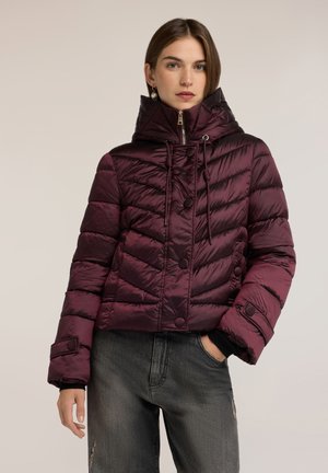 Mujer con chaqueta acolchada de color púrpura oscuro con cuello alto y botones, combinada con vaqueros oscuros desgastados, de pie frente a un fondo liso.
