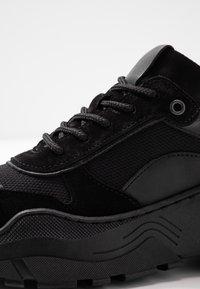 Baskets noires avec une tige en mélange de daim et de mesh, présentant des accents texturés, des lacets ronds, et une semelle en caoutchouc épaisse et rainurée.