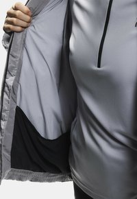 Jasnoszara kurtka z gładkim, błyszczącym wnętrzem i czarnym wykończeniem. Posiada zamek błyskawiczny na pół długości i rękę trzymającą otwartą kurtkę.