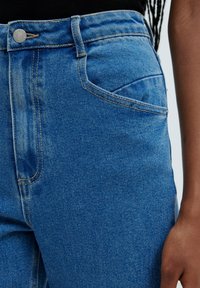 Primer plano de una persona que lleva jeans de mezclilla azules y una camiseta negra, mostrando el bolsillo frontal y parte del brazo derecho contra un fondo claro.