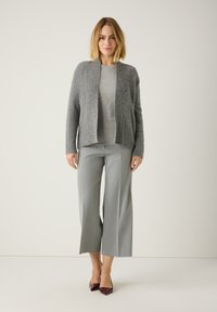 Graue Strickjacke über einem hellgrauen Oberteil, kombiniert mit grauen, weit geschnittenen Hosen und burgunderfarbenen, spitzen Heels. Glatte Texturen sind offensichtlich.