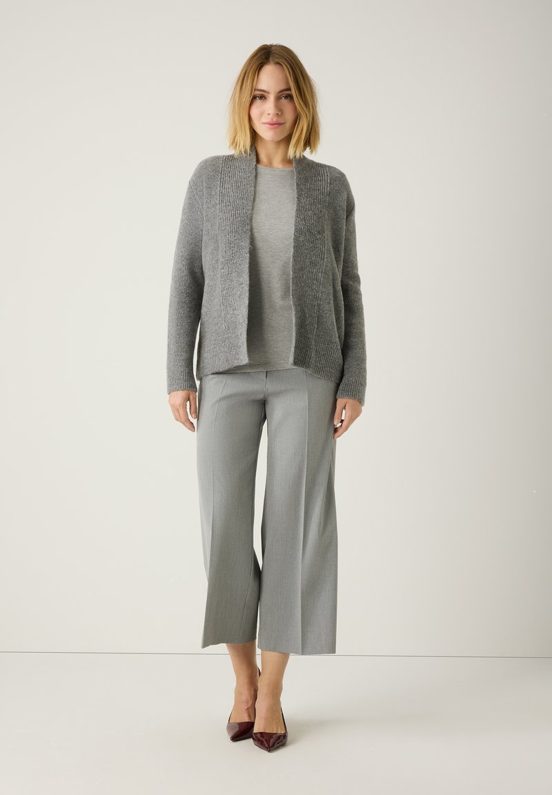 Graue Strickjacke über einem hellgrauen Oberteil, kombiniert mit grauen, weit geschnittenen Hosen und burgunderfarbenen, spitzen Heels. Glatte Texturen sind offensichtlich.