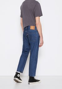 Jeans de denim en azul medio con un corte relajado, que presentan un parche de logo de cuero y dos bolsillos traseros. Combinados con una camiseta gris y zapatillas negras de caña alta.