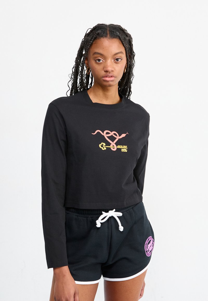 Maglietta nera a maniche lunghe e taglio corto con un design grafico colorato che presenta serpenti e chiavi. Abbinata a pantaloni corti neri con accenti bianchi.
