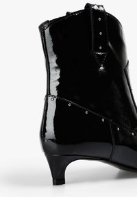 Botín de charol negro con tacón fino, adornado con tachuelas plateadas a lo largo de la costura y un acabado liso y brillante.