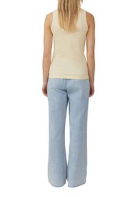 Vrouw met lang blond haar, gekleed in een mouwloos beige top en lichte blauwe wijde jeans, staande met de rug naar voren.