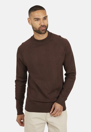 Mann trägt einen braunen gerippten Pullover und hellbeige Hosen, schaut leicht zur Seite mit einem neutralen Ausdruck vor einem weißen Hintergrund.