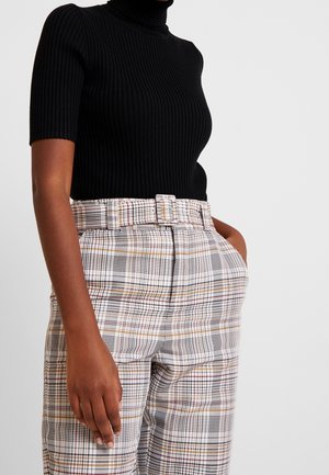 Donna che indossa un dolcevita nero a maniche corte a coste e pantaloni a quadri a vita alta con cintura, con le mani nelle tasche, mostrata dalle spalle fino a metà coscia.