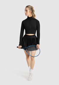 Zwarte cropped sportjack met hoge kraag, gecombineerd met een zwarte geplooide rok. Houdt een blauw-witte tennisracket vast en draagt witte schoenen.