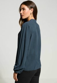 Mørk teal bluse med lange, samlede ærmer og høj hals. Glat, blød tekstur og en afslappet pasform med en buet kant.