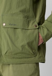 Zelená bunda s texturovanou tkaninou, přední kapsou s klopou a zapínáním na knoflík. Obsahuje vyšívané logo "Barbour" na kapse.