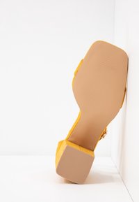 Sandale en daim jaune avec bout carré ouvert et talon bloc, semelle orientée vers l'avant sur un fond d'angle blanc.