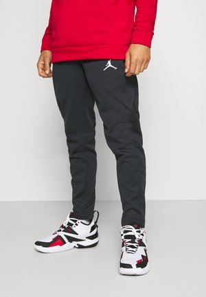 Personne portant un pantalon de sport noir avec un logo Jordan blanc et des chaussures de sport blanches, noires et rouges, debout sur un sol gris.