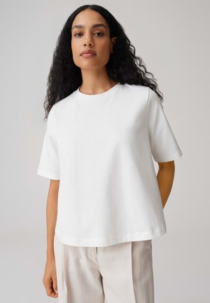 Femme aux longs cheveux bouclés portant un t-shirt blanc uni à manches courtes et un pantalon beige clair sur un fond neutre.
