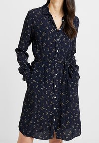 Granatowa sukienka zapinana na guziki z kwiatowym nadrukiem, długimi rękawami i paskiem w talii. Wykonana z lekkiego materiału, z kołnierzykiem i kieszeniami.