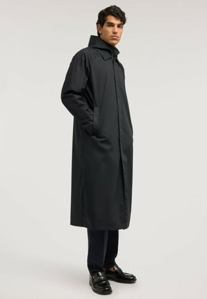 Homme portant un long manteau noir avec une capuche, debout les mains dans les poches, habillé d’un pantalon noir et de chaussures noires.