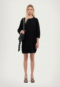 Blonde Frau, die ein schwarzes knielanges Kleid, schwarze Sandalen trägt und eine schwarze Schultertasche mit breiten Trägern und großen Metallösen hält.