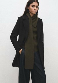 Czarny wełniany płaszcz o dopasowanym kroju, nałożony na ciemnozielony sweter z dzianiny i pasujący do niego szalik, zestawiony z luźnymi ciemnymi spodniami dżinsowymi.