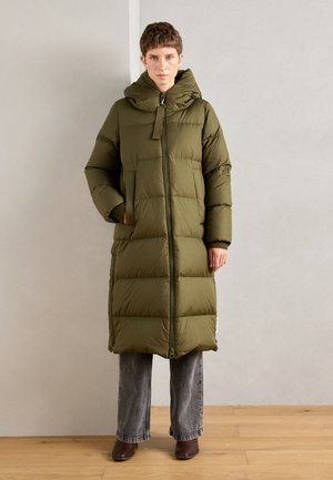 Manteau long rembourré en vert olive, avec une grande capuche, fermeture éclair à l'avant et poches latérales. Porté sur un pantalon en denim gris et des chaussures marron.