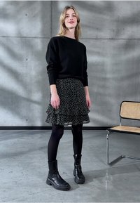 Svart stickad tröja, volangkjol i svart med vita prickar, svarta strumpbyxor och kraftiga svarta ankelstövlar i en minimalistisk betongmiljö.